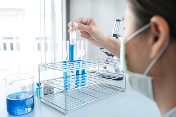 Scientist woman in medical face mask is holding test tube with blue liquid and looking reaction