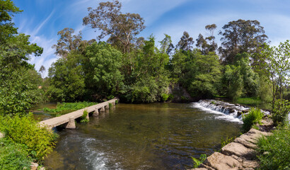 Fototapeta na wymiar Panoramic view of a beautiful little waterfall cascade and an old stone footbridge over the Neiva River in Antas, Esposende, Portugal.