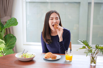 Obraz na płótnie Canvas Young asian woman eating bread with a glass of fresh orange juice and healthy salad while breakfast