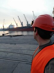 a port worker dressed in work clothes and a helmet, looking at a cargo ship and some cranes at dawn.