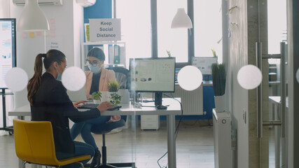 Businesswoman wearing protective face mask working on computer and discussing wiht collegue avout business job. Coworkers keeping social distancing to avoid virus disease during global epidemic