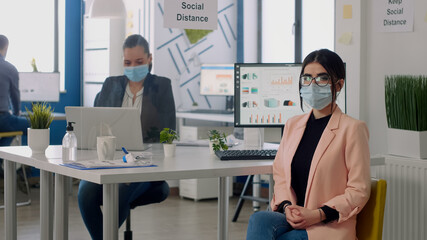Fototapeta premium Portrait of manager with face mask sitting on chair at desk table in nre normal business company office. Teamworkers working in background respecting social distancing during coronavirus pandemic