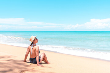 Back view of asian traveler woman in swimsuit and straw hat to sitting on the beach to looking view the blue horizon
