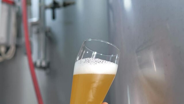 A successful expert Caucasian white brewer with a beard evaluates freshly brewed beer from a beer tank while standing in a beer factory. A modern beer factory. Brewing. Production. Close-up