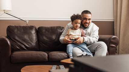 Obraz premium Little boy sitting on the hip of his father and holding joystick. Smiling father and his son playing video games at home.