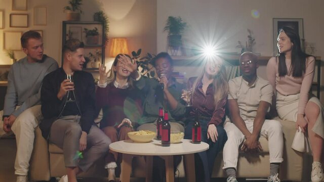 Medium shot of diverse group of young friends sitting on couch and singing karaoke together while celebrating in apartment. Beer bottles and bowls filled with chips are on coffee table