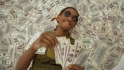 Black man with glasses is lying on a pile of money and waving dollar bills in hands. Concept of wealth and success.