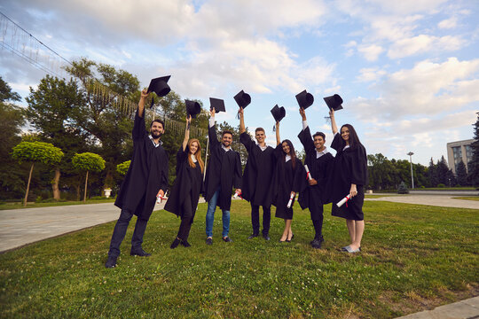 Group of happy university graduates standing on grass with diplomas and holding bonets in raised hands outdoors. Graduation from university concept
