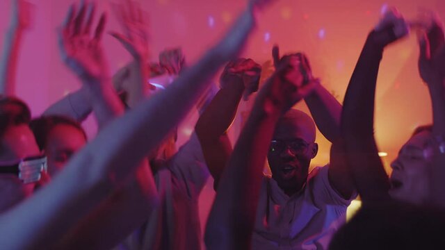 Handheld slowmo shot of diverse group of friends dancing with their arms raised at house party