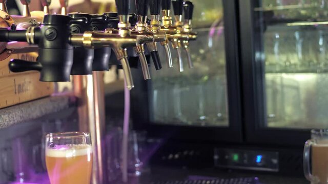 The bartender pours a light beer into a foaming glass. A row of beer steel taps in a dark pub or bar. Taps for draught beer in a modern bar. The bartender pours craft beer at the bar.