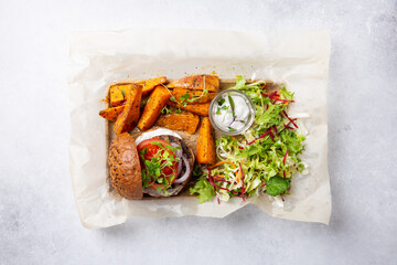Meat free plant based burger served with sweet potato wedges, green mix salad and white sauce on gray table. Healthy vegan or vegetarian food concept. Top view. - obrazy, fototapety, plakaty