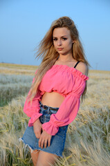 Charming, young woman posing in field of rye. Woman wearing pinky top and jeans skirt.