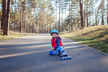 Sad kid in sport helmet sitting on road during riding on roller skates at park, active family weekend, spring outdoors activity, healthy lifestyle