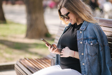 Pregnat woman sits on bench in the park and using phone. Happy female have rest outdoor during the walks