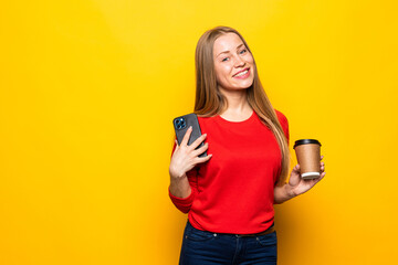 Beautiful young woman drinking coffee using mobile phone posing isolated over yellow wall background