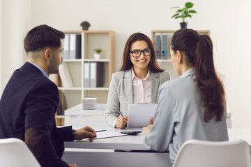 Friendly smiling professional insurance expert or loan broker in glasses meeting clients sitting at desk at her office. Young couple consulting bank manager and asking advice on money and finance