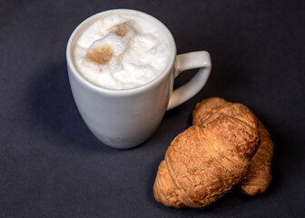 Latte with milk froth in a white cup on a dark background. Coffee and croissants. Close-up