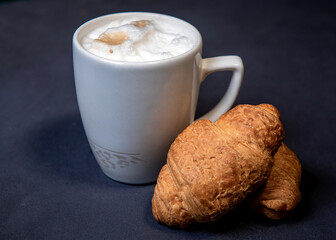 Latte with milk froth in a white cup on a dark background. Coffee and croissants. Close-up