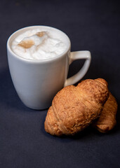 Latte with milk froth in a white cup on a dark background. Coffee and croissants. Close-up