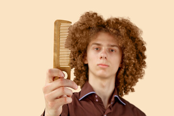 A long-haired curly-haired guy in a brown shirt on a beige background uses a wooden comb. Emotions before a haircut in a hairdresser