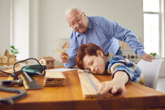 Grandfather And Grandson Fixing Things In Workshop. Skillful Carpenter Teaching Little Child New Handwork Skills. Old Man With Teen Boy Measuring Wood Plank To Make Wooden Toys Or Do Home Repairs