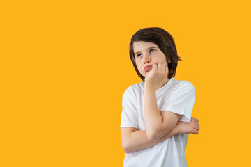 Portrait of a young boy of nine years old wearing white t-shirt, pressing his hand to his face, thinking seriously and looking up. Pensive child isolated on yellow background, copy space.