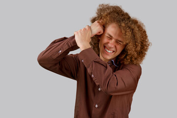 A long-haired curly-haired guy in a brown shirt on a gray background uses a wooden comb. Emotions before a haircut in a hairdresser. Pain from combing