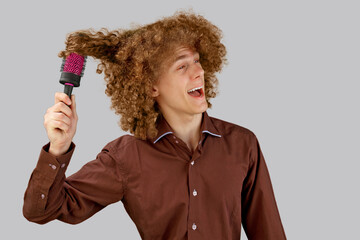 A long-haired curly-haired guy in a brown shirt on a gray background uses a metal round comb. Emotions before a haircut in a hairdresser. Pain from combing