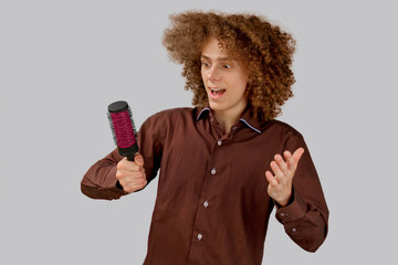 A long-haired curly-haired guy in a brown shirt on a gray background uses a metal round comb. Emotions before a haircut in a hairdresser