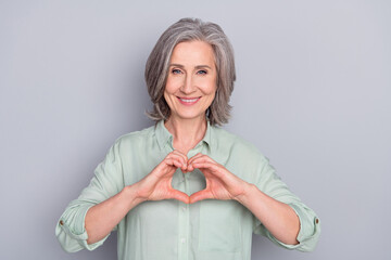 Photo of mature smiling beautiful cheerful lovely woman showing heart sign gesture isolated on grey...