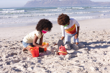 African american mother with son wearing face mask playing with sand at the beach