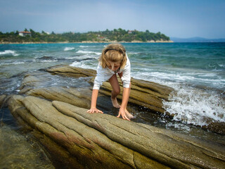 A barefoot blond girl in the white sweater walks along the rocky shore  surrounded by turquoise sea and beautiful nature. 