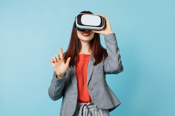 Positive human emotions, facial expressions. Pleasant young woman in bright trendy formal clothes, wearing a virtual reality headset on a blue studio background, pointing up.