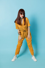 Fototapeta na wymiar Full length view of young gorgeous red haired lady in bright yellow trendy overalls and sunglasses, posing to camera on isolated blue color background. Summer concept, studio shot.