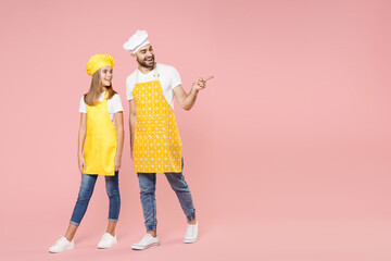 Full length body of teen girl dad man father's helper chef cook confectioner baker in yellow apron toque cap uniform walking going point finger away isolated on pastel pink background studio portrait.