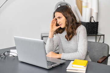 Worried businesswoman sits at the desk in front of laptop, looks away and lost in thoughts, upset female employee concerned about project, late with deadline, has not inspiration