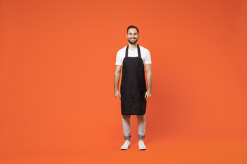 Fototapeta na wymiar Full length young fun man 20s barista bartender barman employee in black apron white t-shirt work in coffee shop looking camera isolated on orange background studio . Small business startup concept.