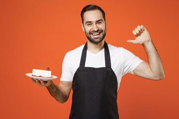 Young man barista bartender barman employee in black apron white t-shirt work coffee shop point finger on cake dessert point thumb finger himself isolated on orange background Small business startup.