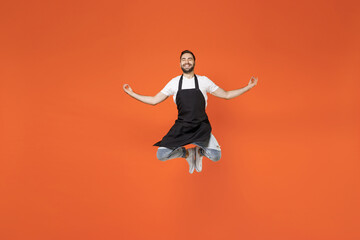Full length young man 20s barista bartender barman employee in black apron white t-shirt work in coffee shop jump high do yoga om gesture isolated on orange background. Small business startup concept.