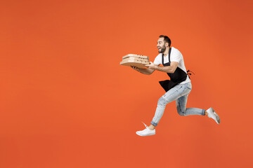Fototapeta na wymiar Full length side view young man barista barman employee in apron white t-shirt work in coffee shop jump hold italian pizza in cardboard flatbox run isolated on orange background Small business startup
