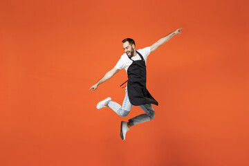 Fototapeta na wymiar Full length young fun man barista bartender barman employee in black apron white t-shirt work in coffee shop jump high outstretched hands isolated on orange background. Small business startup concept.