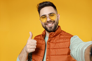 Close up young smiling happy fun caucasian man 20s in orange vest mint sweatshirt glasses doing selfie shot on mobile phone show thumbs up like gesture isolated on yellow background studio portrait