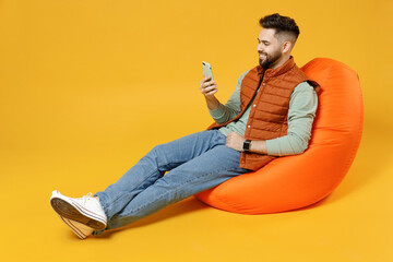 Full length young smiling happy fun caucasian man 20s years old wear orange vest mint sweatshirt sitting in beanbag bag chair hold mobile cell phone chat isolated on yellow background studio portrait.