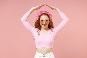 Young fun smiling happy woman 20s in rose clothes bandana glasses hold folded hands above head like house roof, stay home isolated on pastel pink background studio portrait. People lifestyle concept