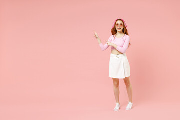 Full length young smiling trendy woman in rose clothes bandana glasses point index finger aside on copy space area mck up isolated on pastel pink background studio portrait People lifestyle concept.