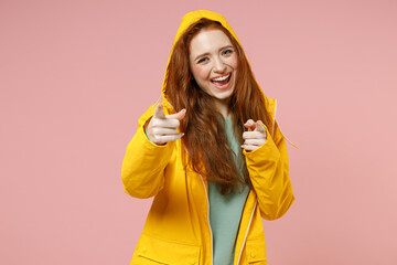Redhead young woman in yellow waterproof raincoat outerwear put on hood point index finger camera on you motivate isolated on pastel pink background studio Outdoors lifestyle wet fall season concept