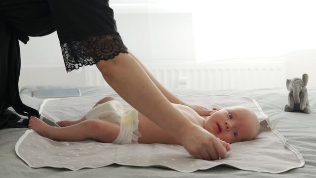 Mother with baby boy doing exercise simple exercises to make baby bones and muscles stronger, newborn infant lying on bed against the window. High quality 4k footage