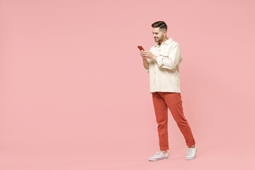 Fototapeta na wymiar Full length young smiling happy fun fashionable friendly caucasian man in jacket white t-shirt using mobile cell phone chat online in social network walk go isolated on pastel pink background studio.
