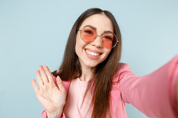 Close up young happy fun friendly caucasian woman 20s in pastel pink clothes glasses doing selfie shot on mobile phone waving hand hello greeting talk by video call isolated on blue background studio