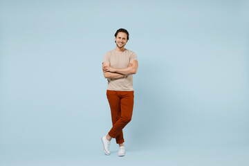 Full length young smiling happy positive friendly caucasian man 20s wearing casual basic beige t-shirt looking camera hold hands crossed folded isolated on pastel blue color background studio portrait